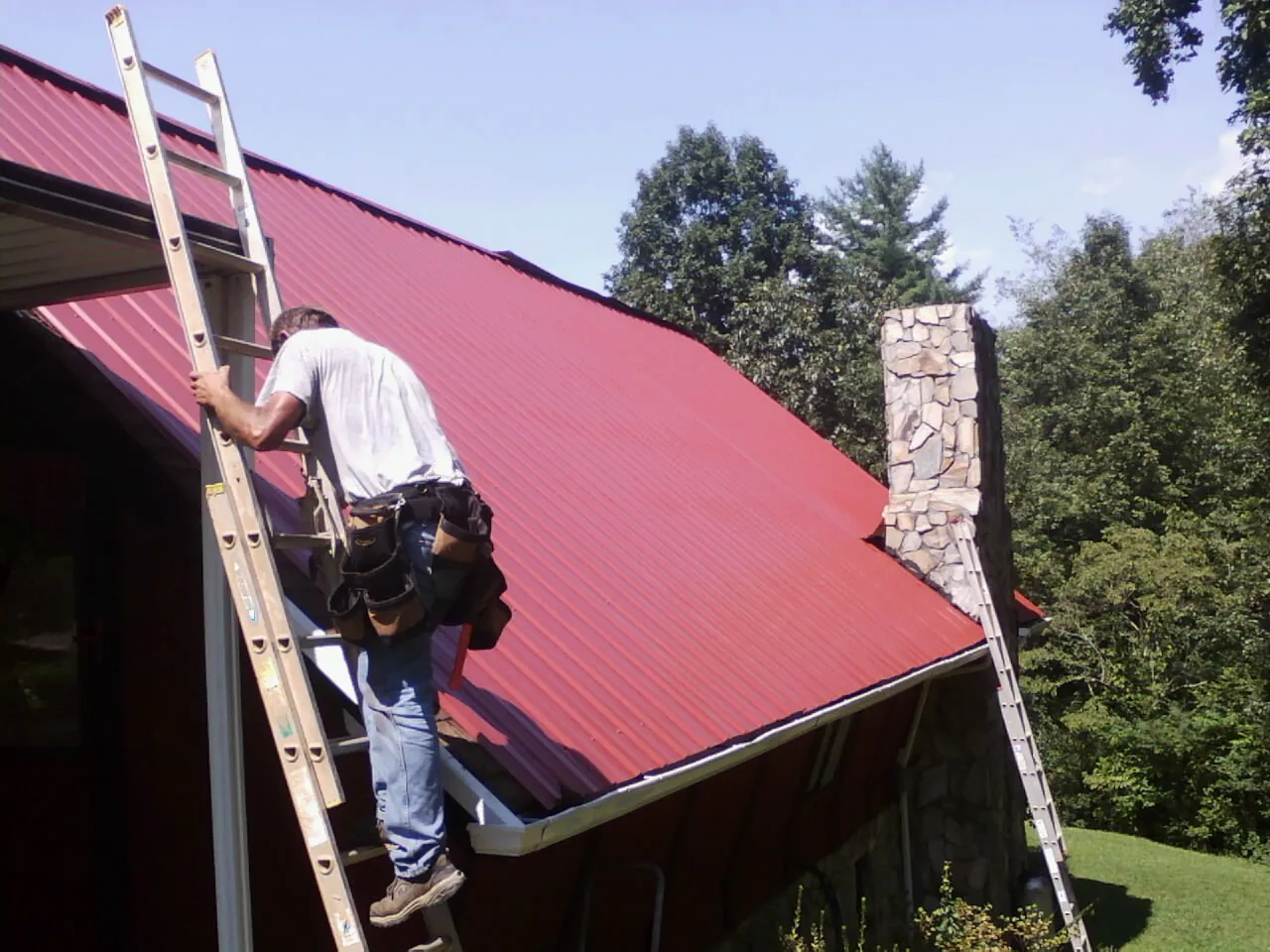 Roofer on a ladder beside a red metal roof and stone chimney — Soffit & Fascia Repair by Roofing Brothers in Roulette